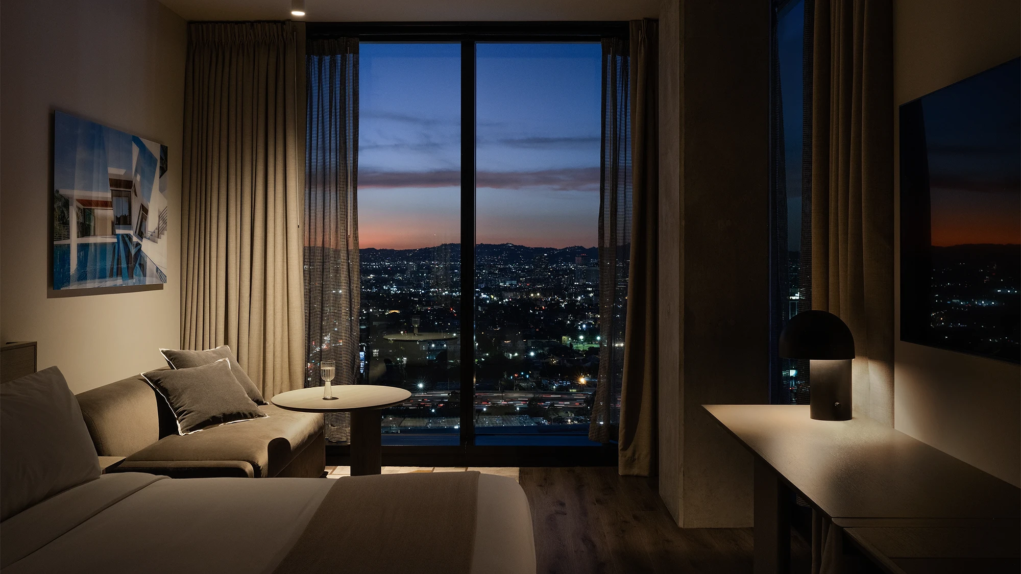 Stylish hotel room looking out at the Los Angeles sunset, featuring a bed, desk, and well-designed lighting fixtures