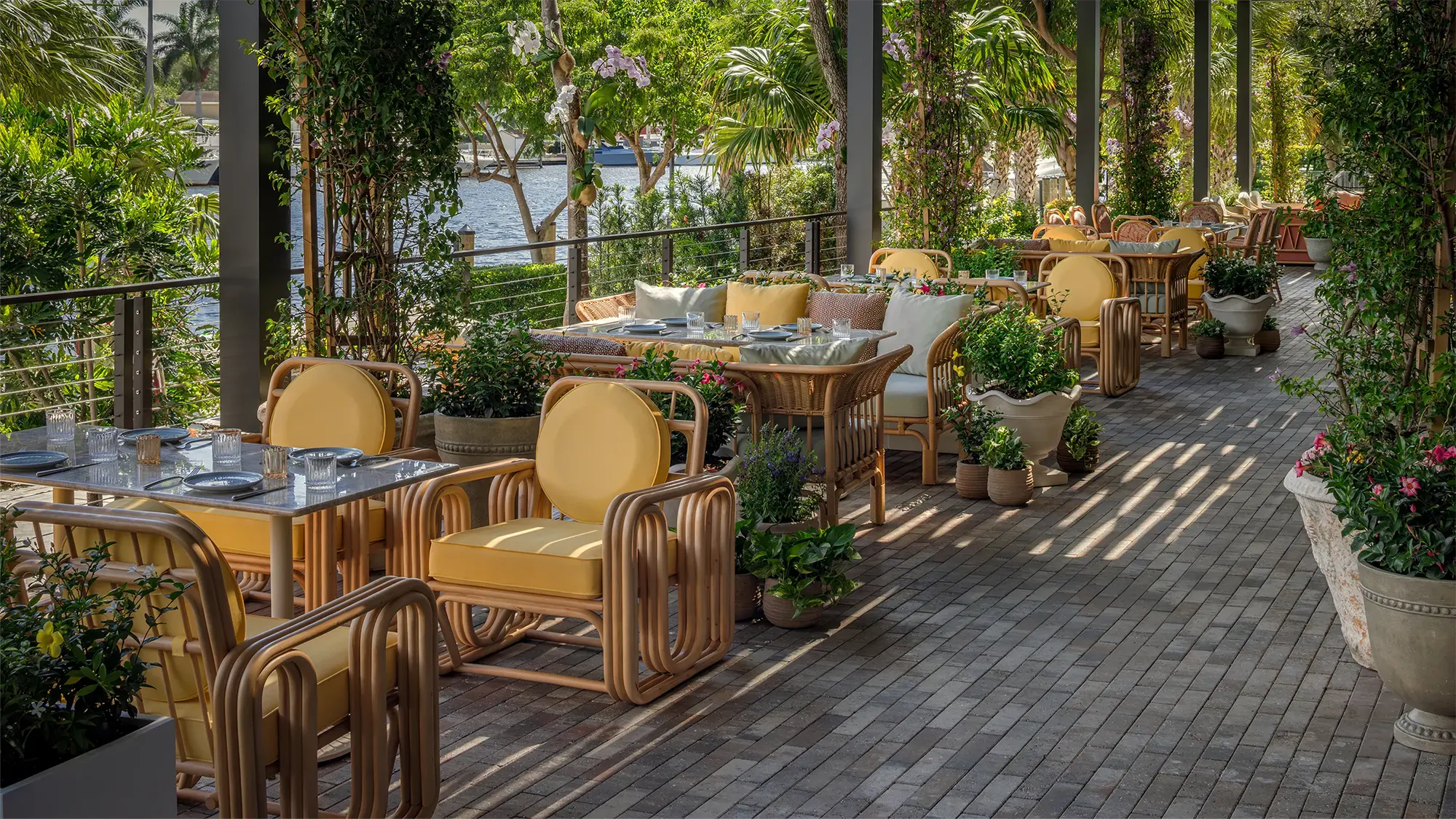 glass top outdoor tables with two sets of wooden chairs with yellow cushions on a tile terrace covered with plants overlooking a river on a sunny day