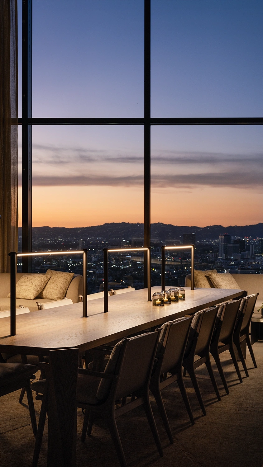 Wooden tables with elegantly designed chairs, illuminated by three stylish lights, set against a large window at sunset.