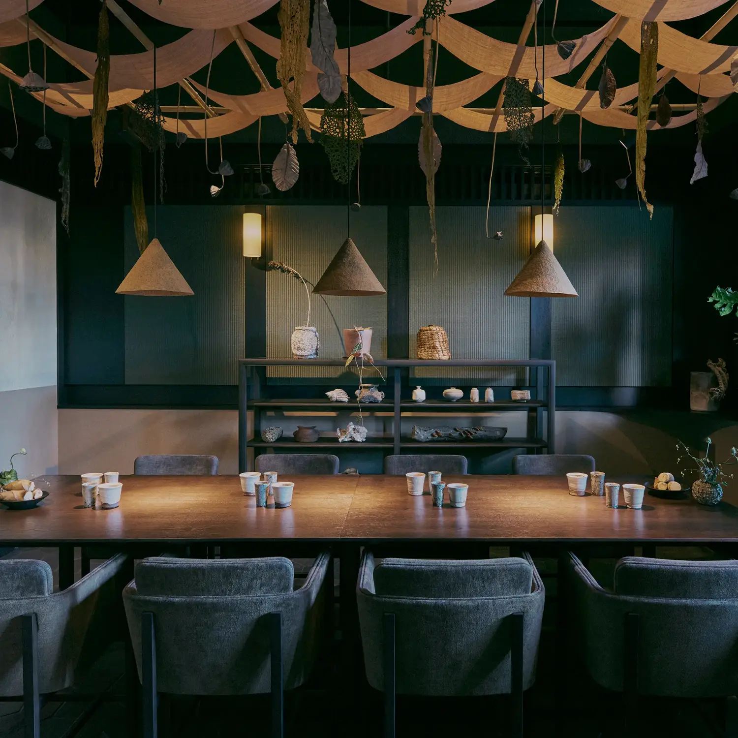 A large wooden dining table set with glasses for each chair, lit by three hat-like lighting fixtures