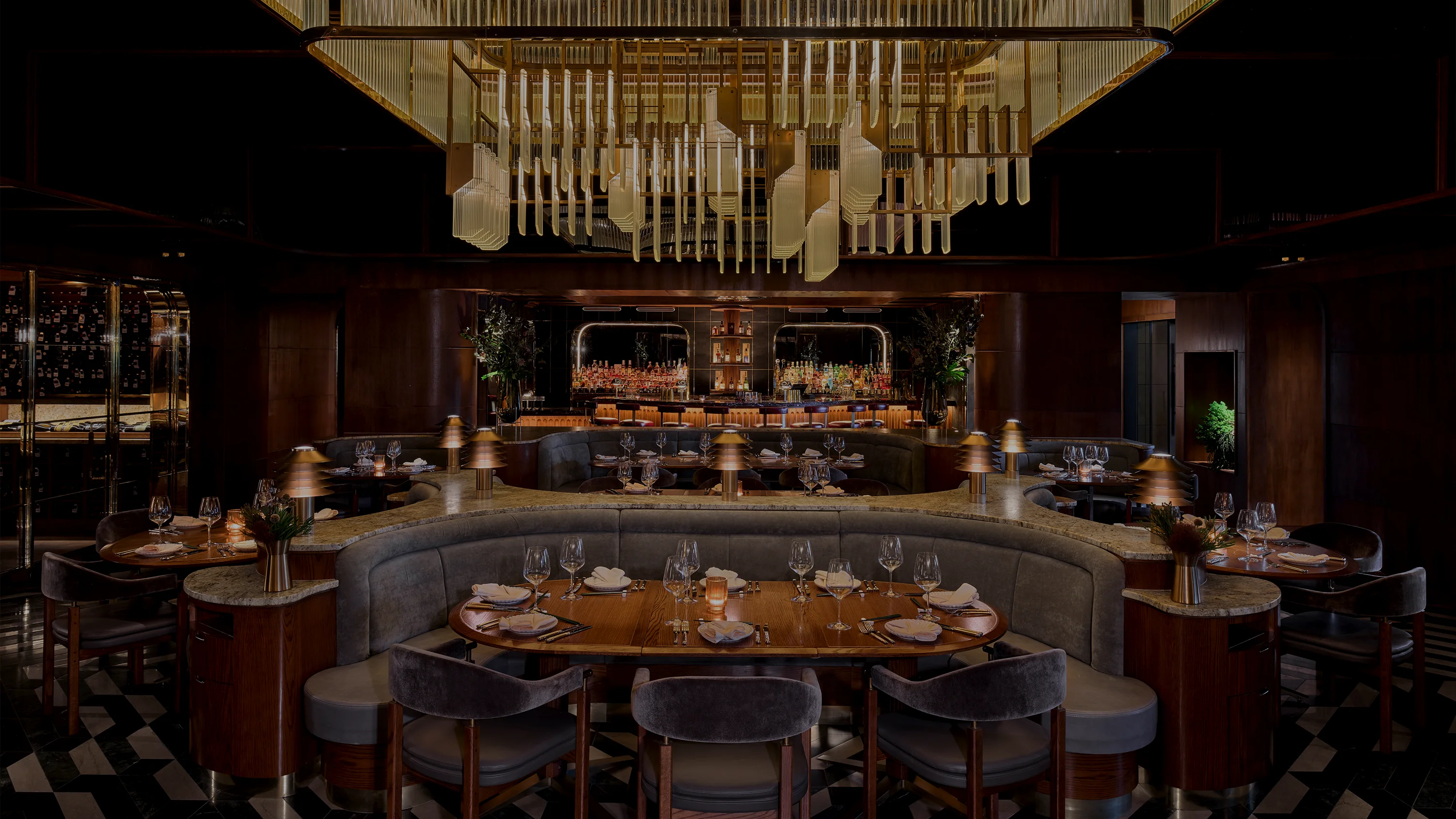 A restaurant seating area with three dinner-ready wooden tables around a center upholstered couch-style seating lit by an elegant chandelier