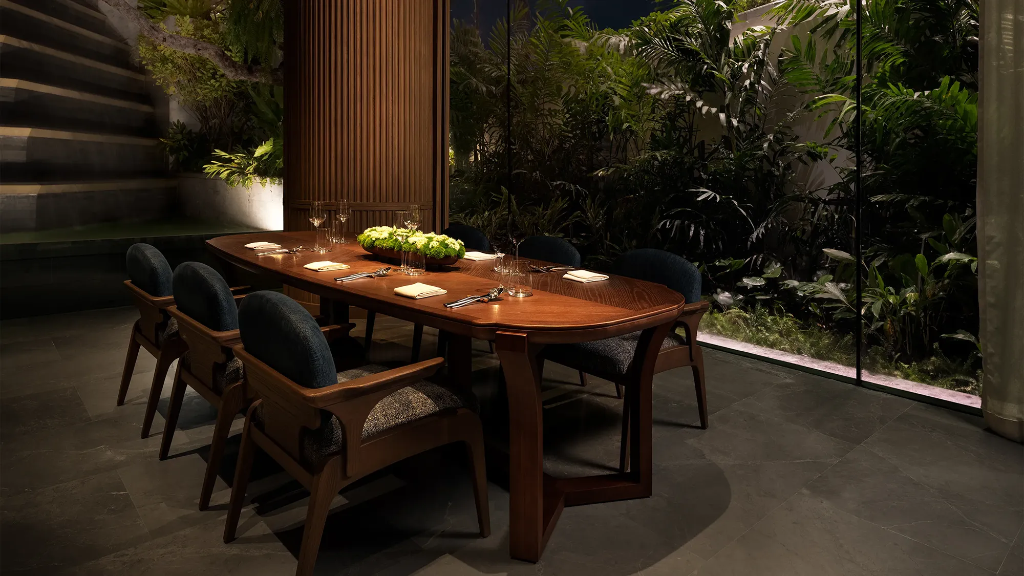 Private dining seating area with a large wooden table, lined by rows of three wooden chairs in front of a window looking into a small indoor tropical garden