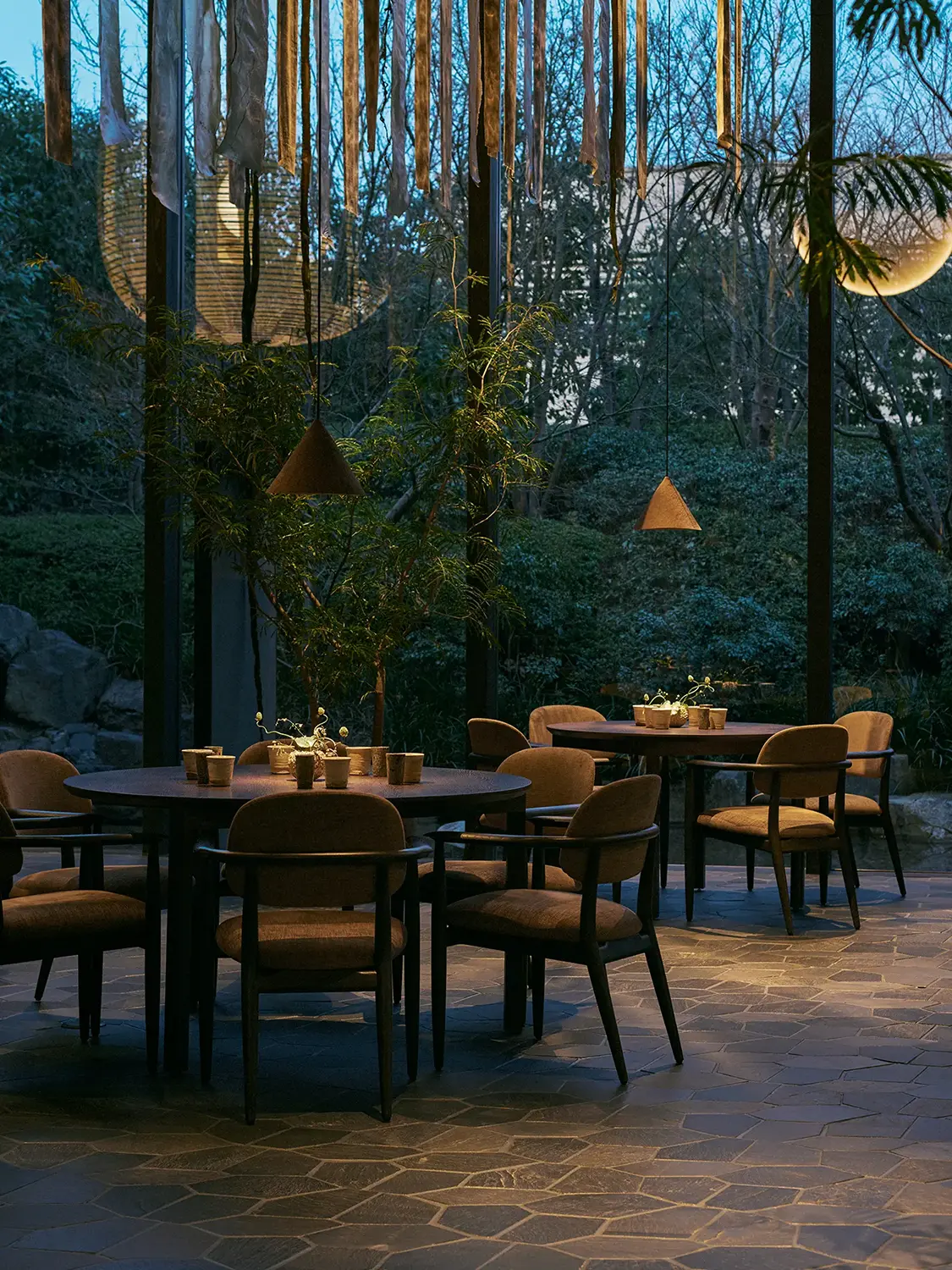 A curated green garden outside of floor-to-ceiling windows behind a dimly lit restaurant dining area