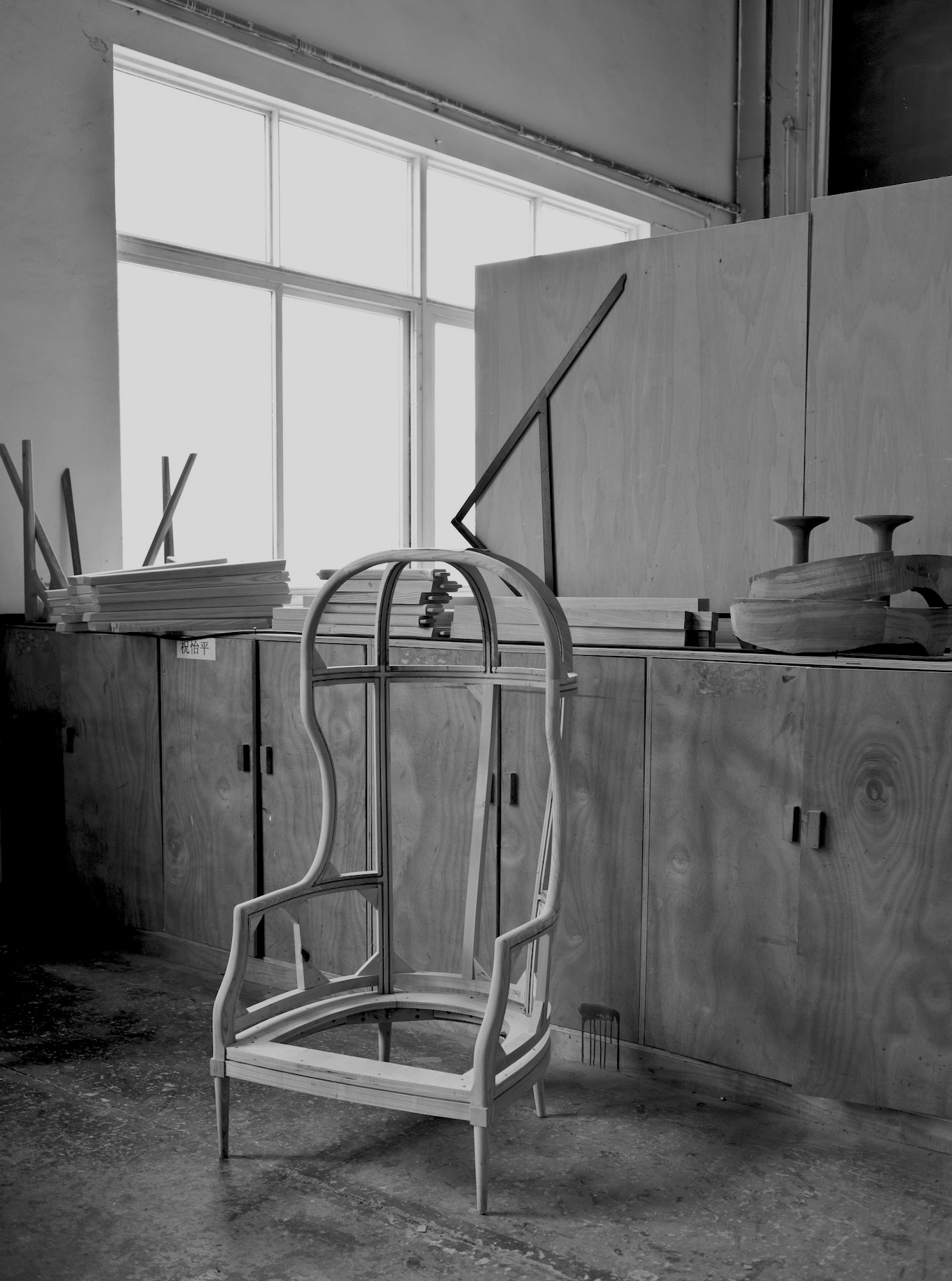 A wooden chair partially manufactured before upholstery stages, sitting in front of a row of cabinents with partially built furniture stacked on top backlit by a  metal framed window